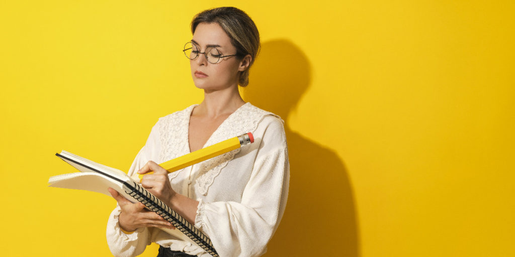 Femme sérieuse portant des lunettes tenant un grand crayon et un carnet sur fond jaune